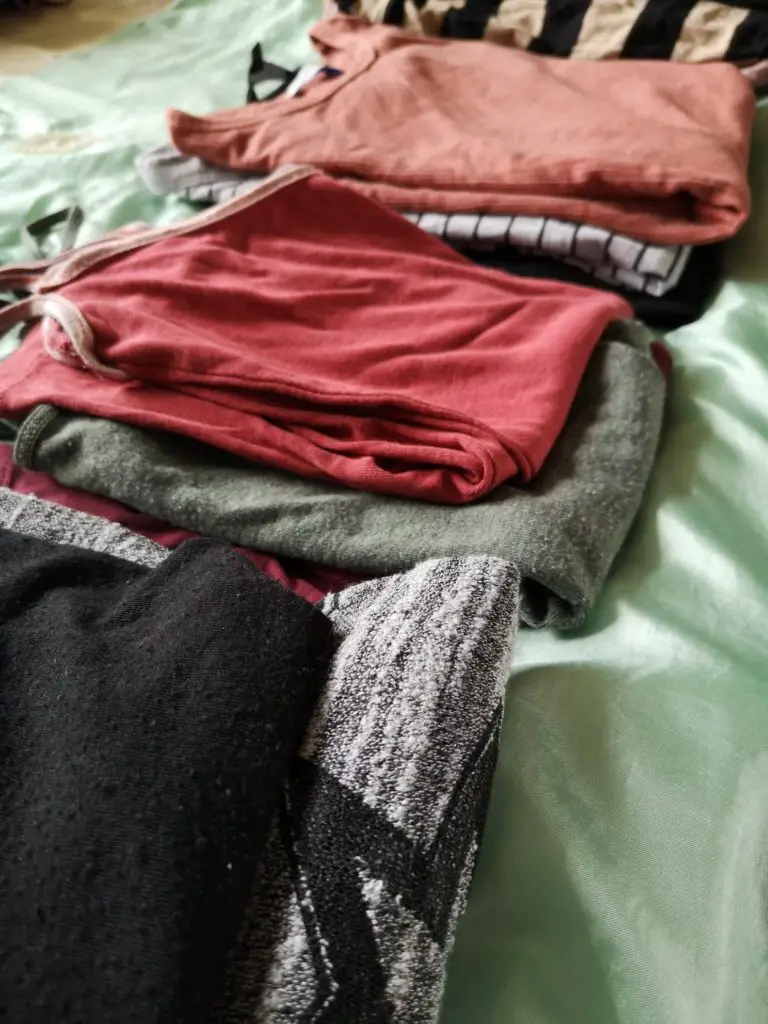 Several piles of tops in Autumnal colours, laid out on a bed.