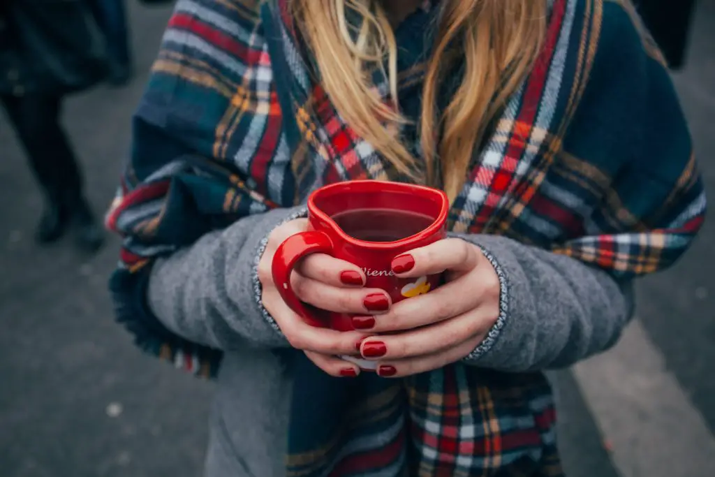 A woman wearing a tartan shawl over a grey jumper. Her nails are painted red and she is holding a red mug containing a hot drink.