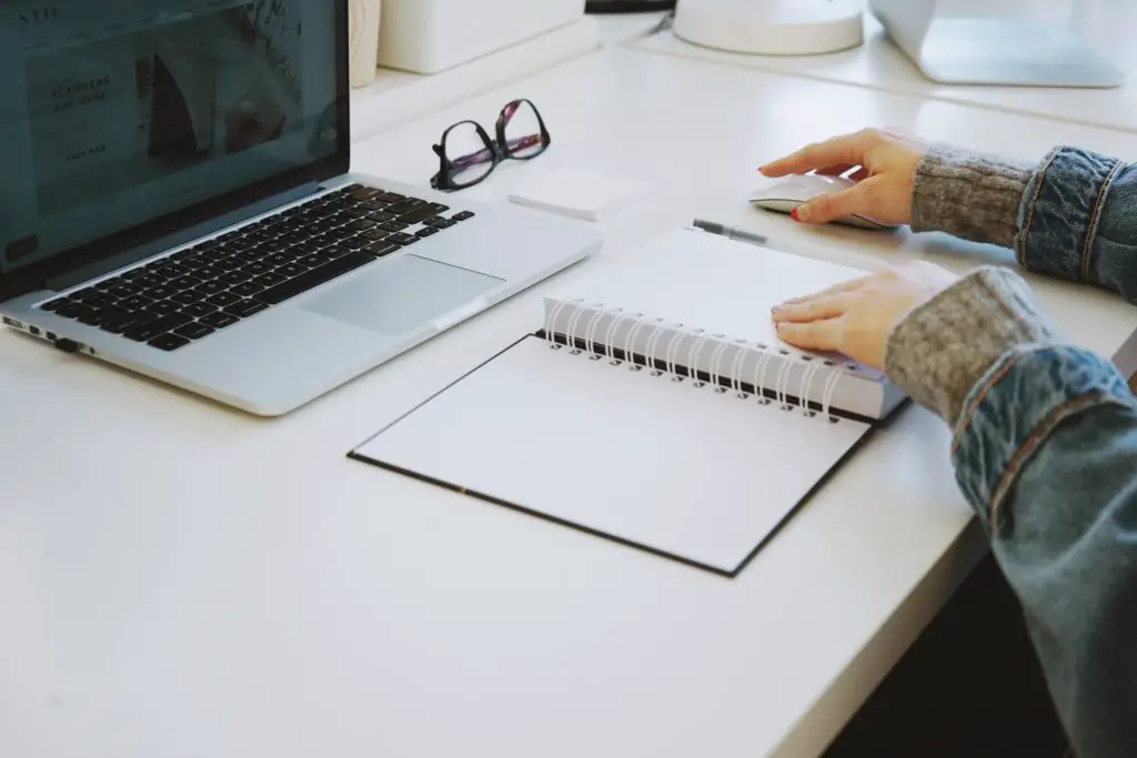 A planner in front of a laptop. Hands resting on the mouse and planner and a pair of glasses on the desk.