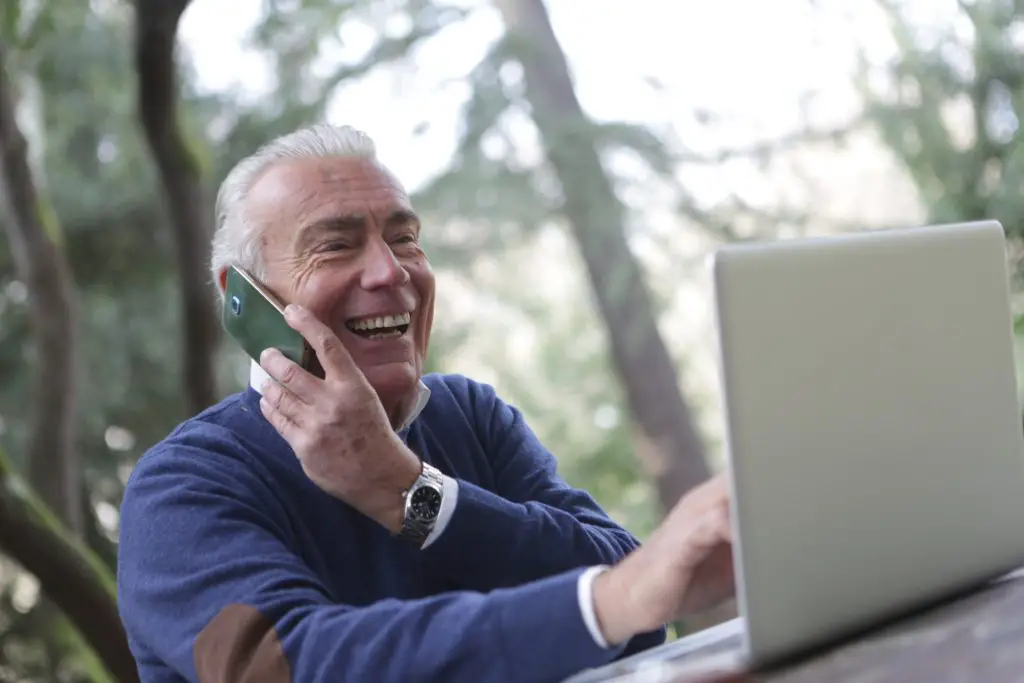 A man holding a phone to his ear whilst on his laptop. He is outdoors and smiling.