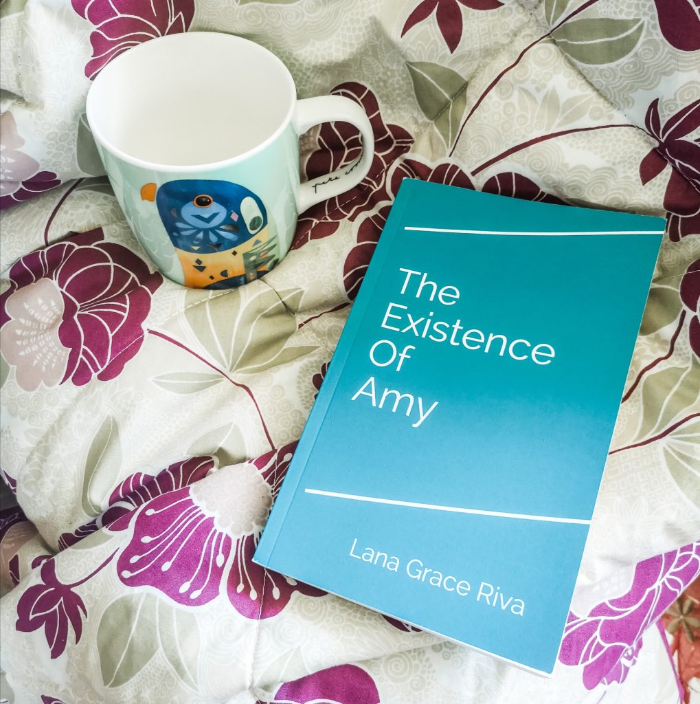 Paperback copy of The Existence Of Amy, laid on a flowery throw. A mug is placed next to it.