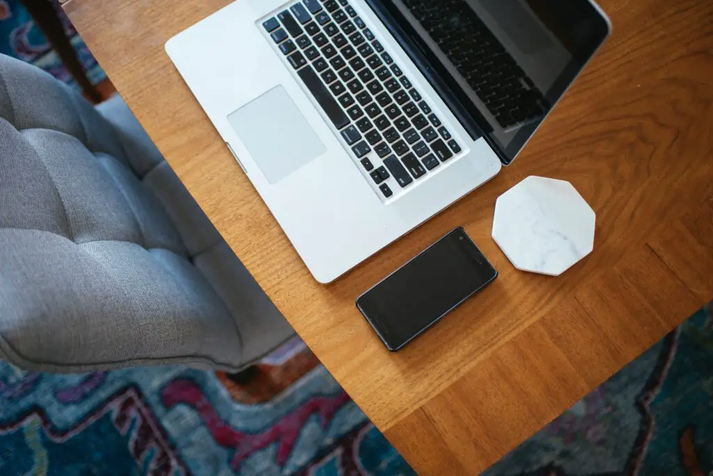 A silver Macbook on a brown wooden desk. There is a phone and white marble coaster next to it. A grey chair is tucked under the desk and there is a blue patterned carpet.