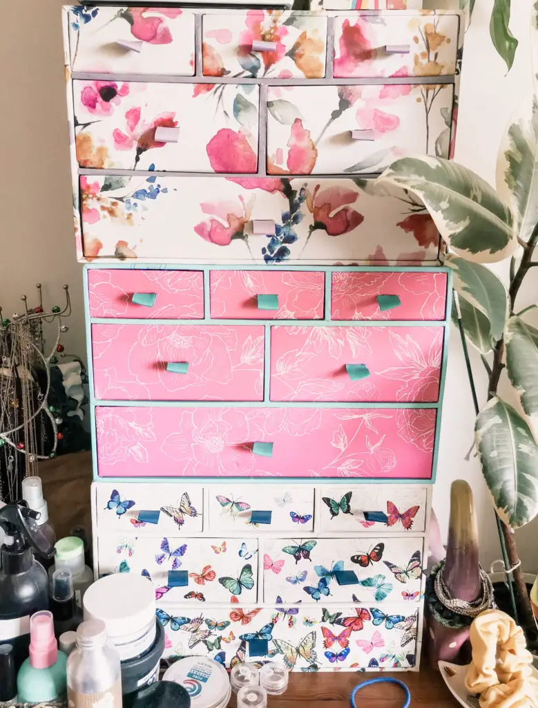 3 colourful sets of mini drawers, stacked on top of each other. The drawers have floral and butterfly patterns on them. They are surrounded by beauty products and a houseplant.