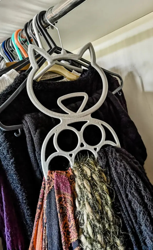 Various scarves hanging on a grey, cat-shaped scarf hanger. The scarf hanger is hanging in a wardrobe with other clothes hanging behind it.