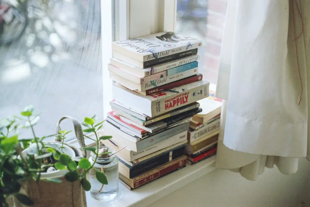 A stack of books on a windowsill with houseplants next to it.