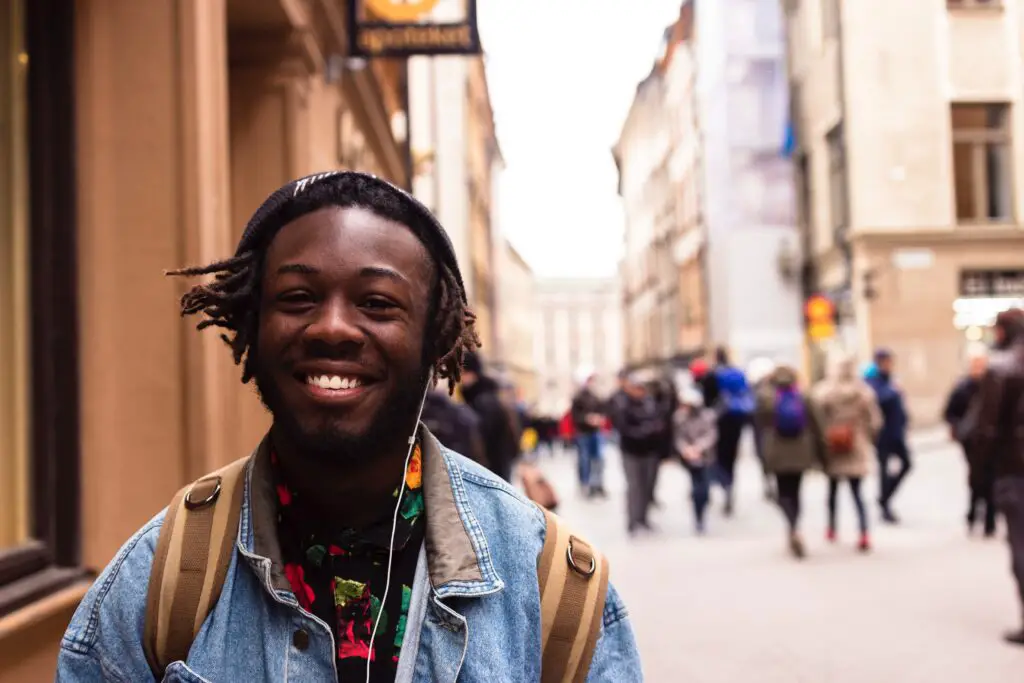 A man wearing headphones and smiling as he walks down the street. There are other people walking in the background.