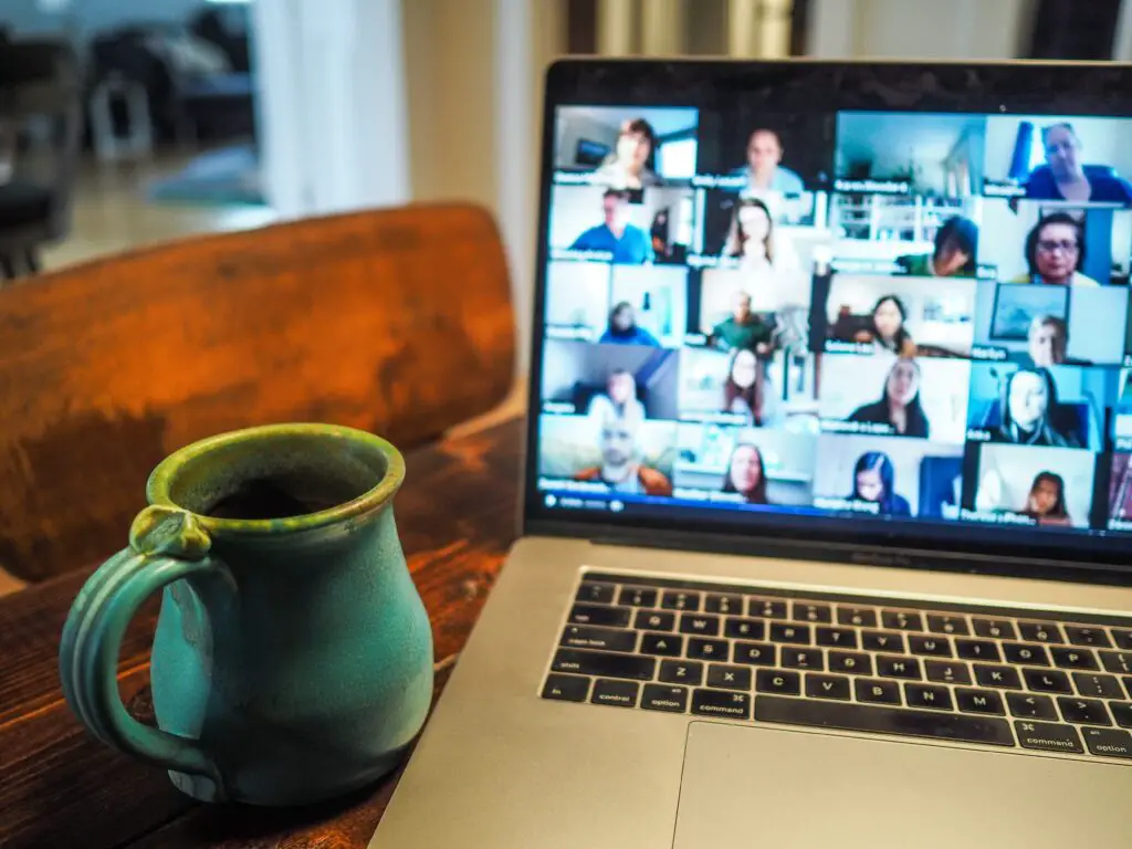 A laptop screen with a group video call occurring - many faces are on the screen. There is a green mug next to the laptop.
