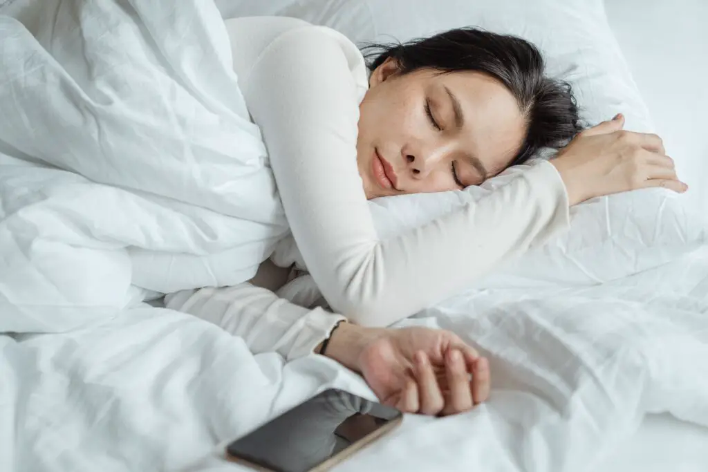 A woman asleep in bed with her phone laying next to her outstretched hand.