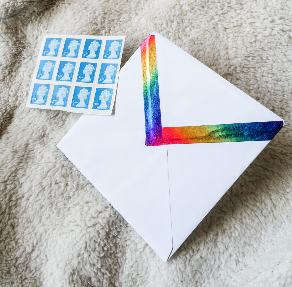 A white envelope that has been sealed with rainbow coloured washi tape. Next to it is a sheet of second class stamps.