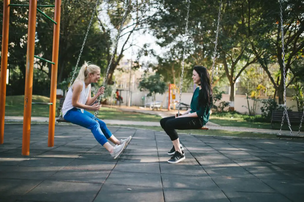 Two women sitting on swings at a park, talking and laughing.