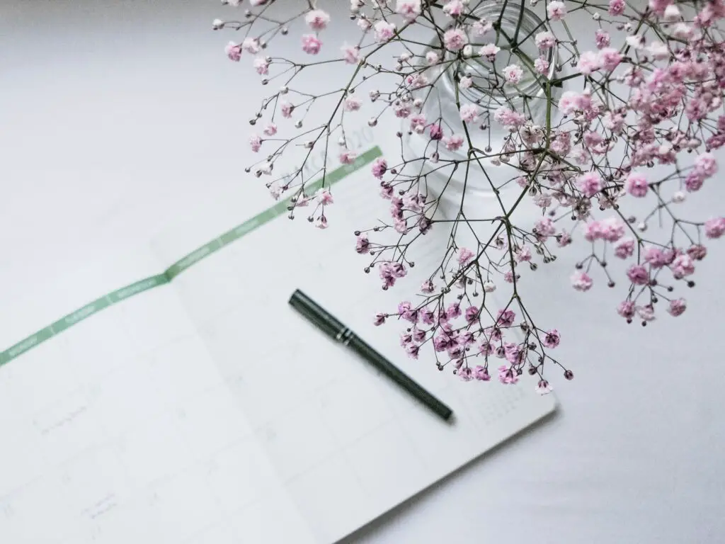 A planner and pen with a vase of delicate pink flowers next to it.