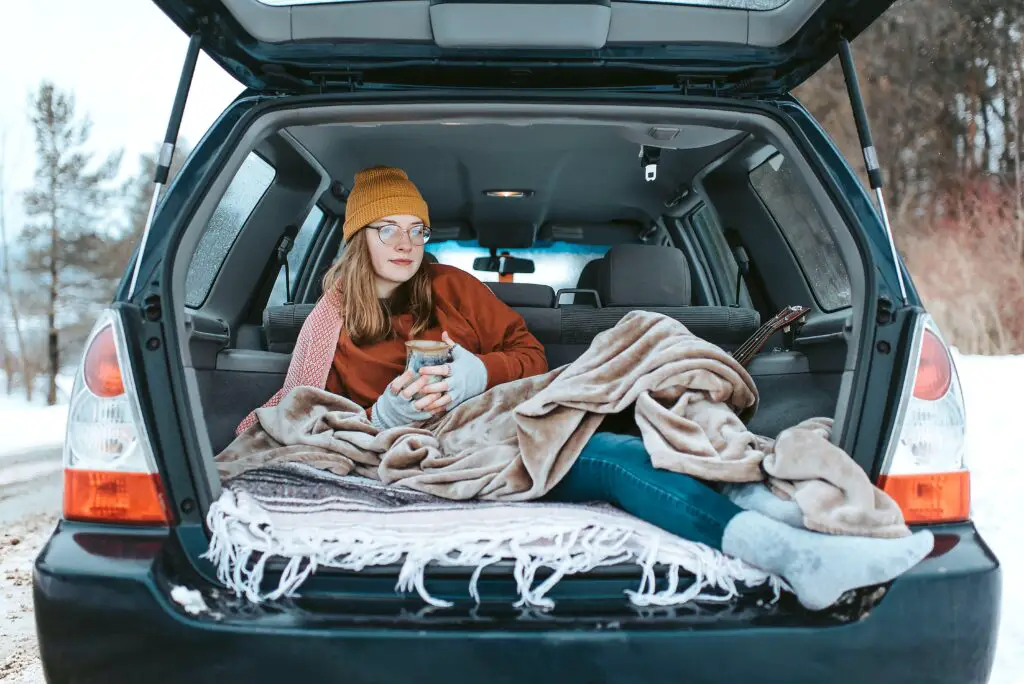 A woman sat in the back of a car, wrapped up in warm clothes and blankets and drinking a hot drink from a mug.