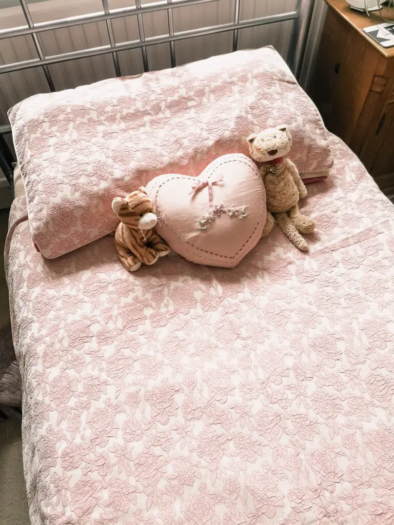 A bed with pink floral duvet cover and pillow. There is a pink, heart-shaped cushion on the bed as well as two cat soft toys.