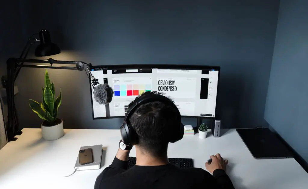 A behind view of a man working on graphic design on a computer. He is wearing a headset and has a microphone beside him.