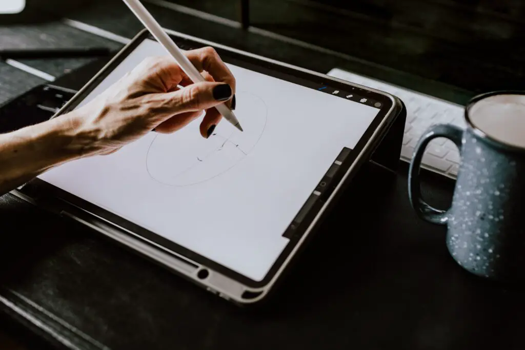 A close-up view of a hand holding a digital pencil and drawing a face on a tablet. There is a mug of coffee next to the tablet, and a keyboard in the background.