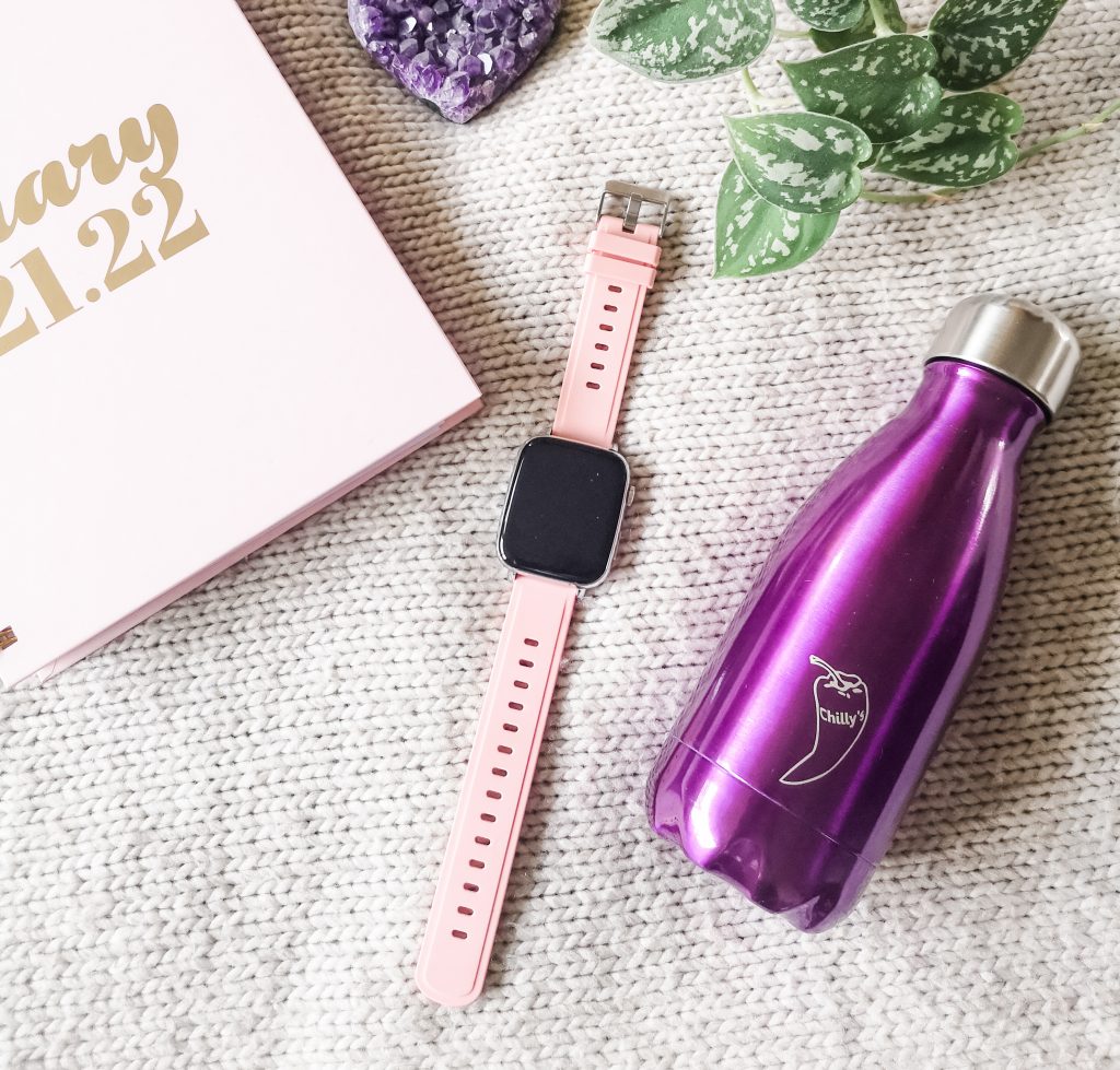 The Misirun Smart Watch laid out among a purple water bottle, pink diary, purple crystal heart and green plant
