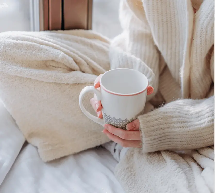 A woman sitting cross-legged on a bed, wearing warm, cosy pyjamas and holding a mug.