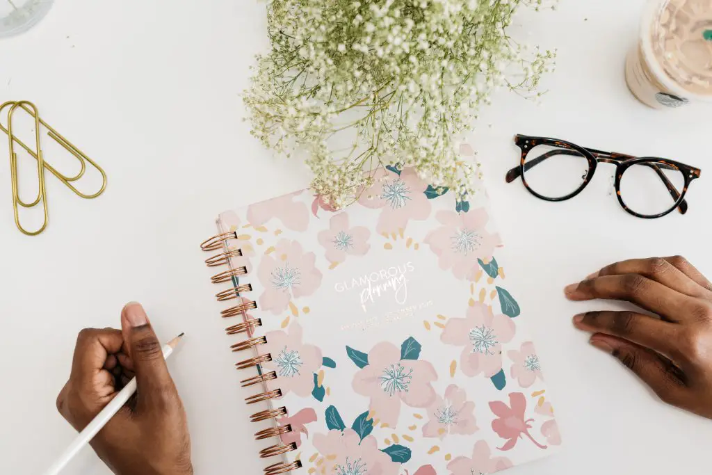 A flat lay of a floral notebook with the words 'glamorous planning' on the front. There is a pair of hands, one of which is holding a pencil. Also, a pair of glasses, two paperclips, a Starbucks cup and some white flowers.