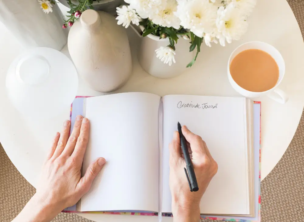 A birds-eye view of somebody's hands holding open a journal and writing the words 'Gratitude Journal' in it. Next to it on the table are a cup of tea and several vases, some of which have white flowers in them.