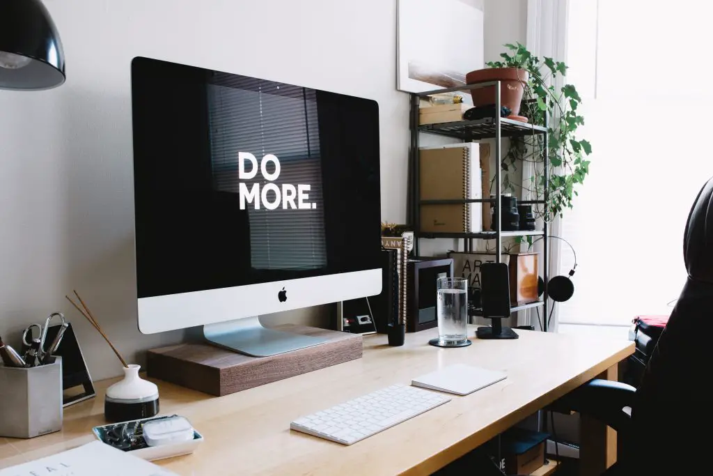 A desk with an apple desktop monitor showing the words 'Do More.' Surrounding the monitor are shelves, books, stationery and plants.