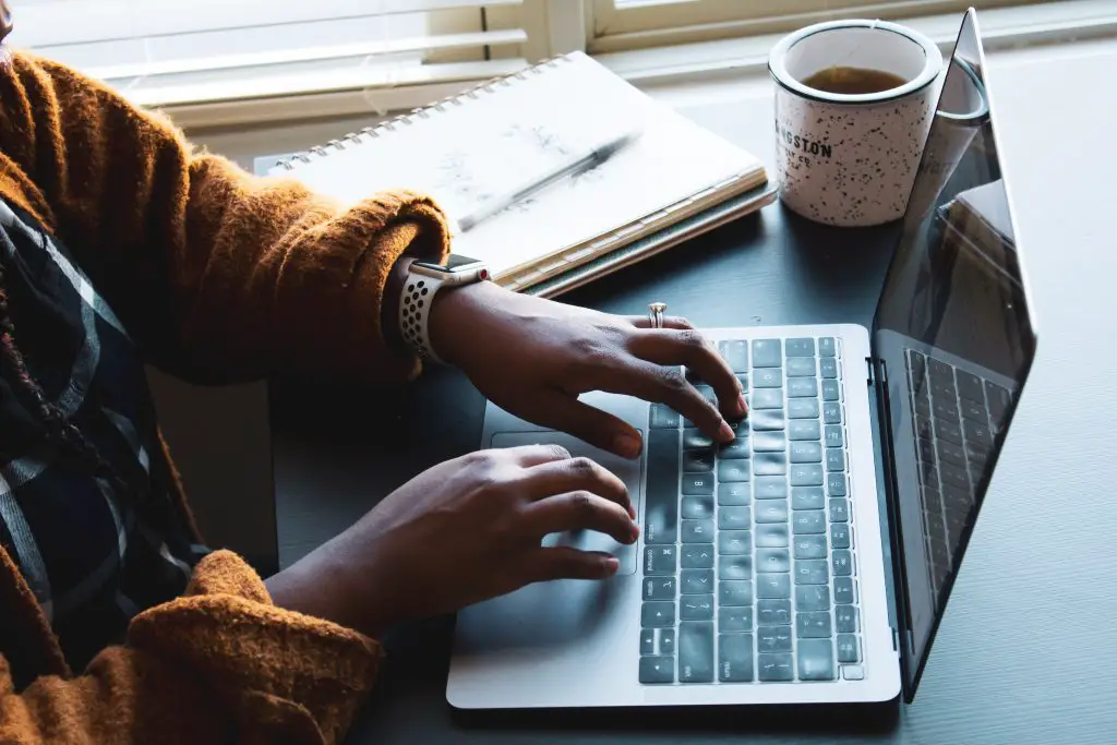 A woman typing on a laptop. There is a notebook and pen and a cup of tea next to her hands on the keyboard.