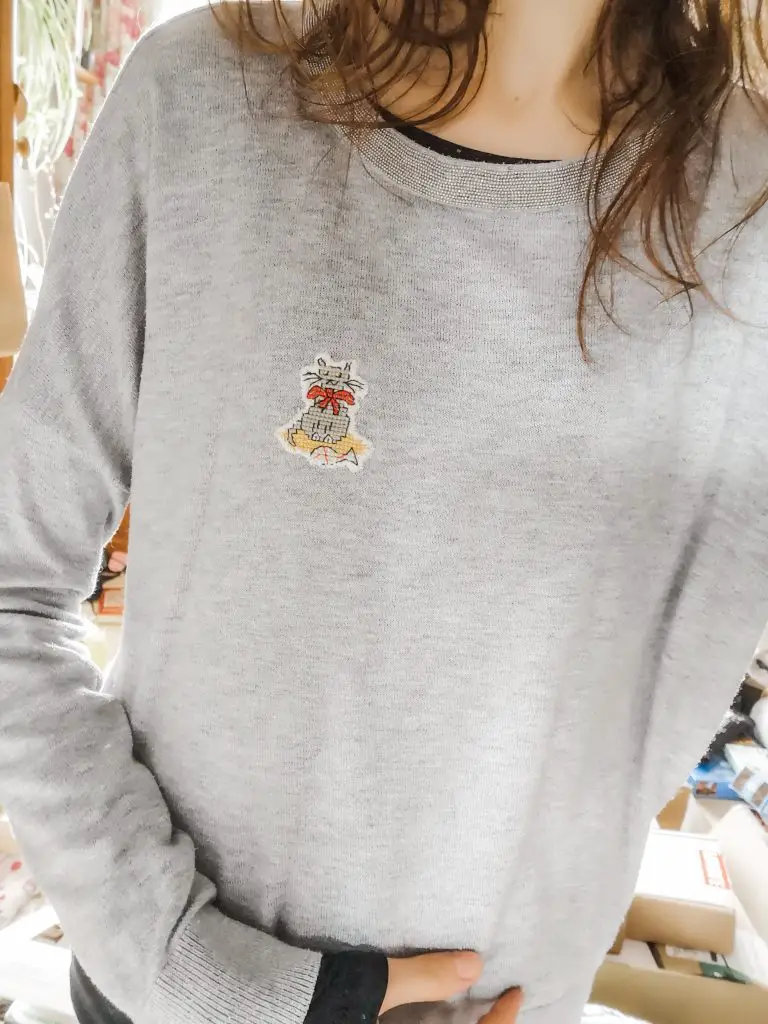 A woman wearing a light grey jumper with cross-stitch cat sewn onto the front.