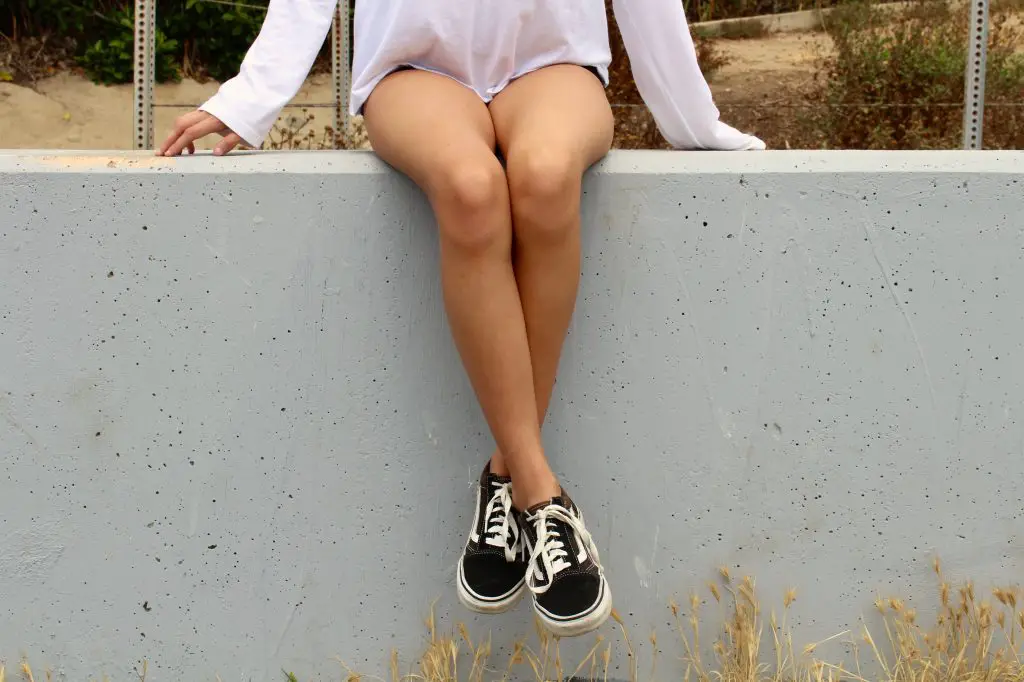 A woman sitting on a wall with her legs dangling down. She is wearing a white top and black trainers and her bare legs are crossed at the ankles.