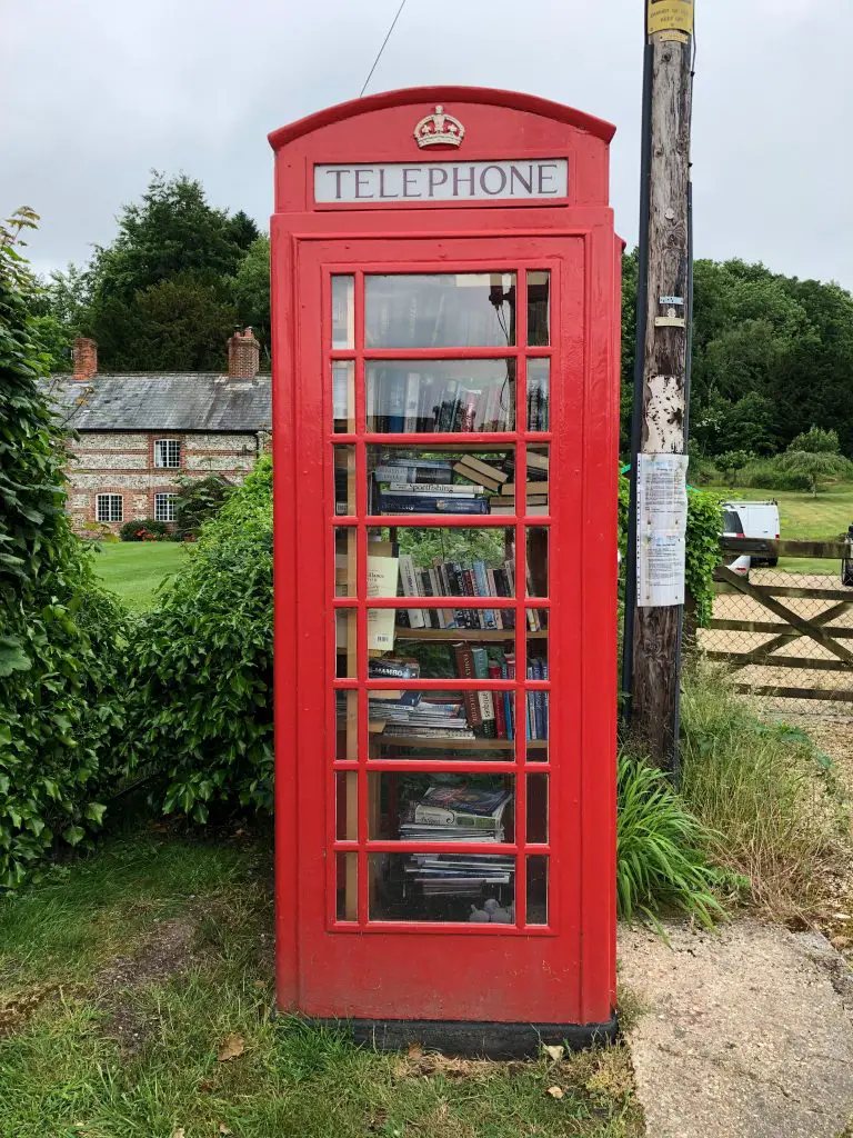 A red telephone box in the countryside, filled with shelves of books.