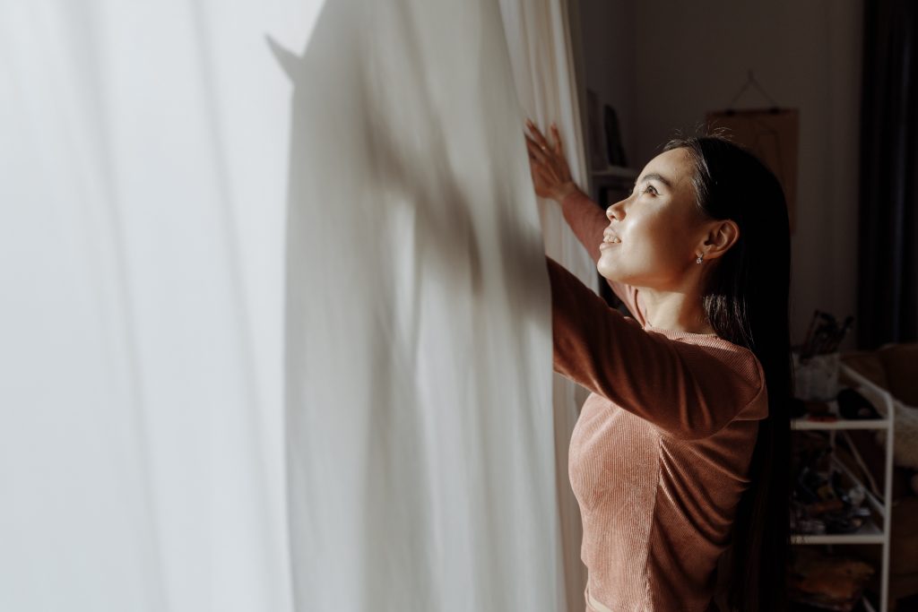 A woman opening some white curtains and looking upwards out of the window.