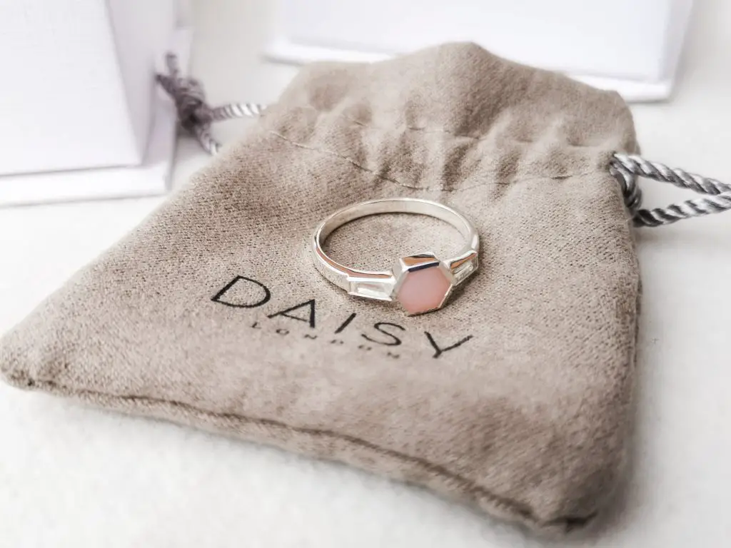 A silver ring with a pink opal gemstone, resting on top of a small Daisy London drawstring bag.