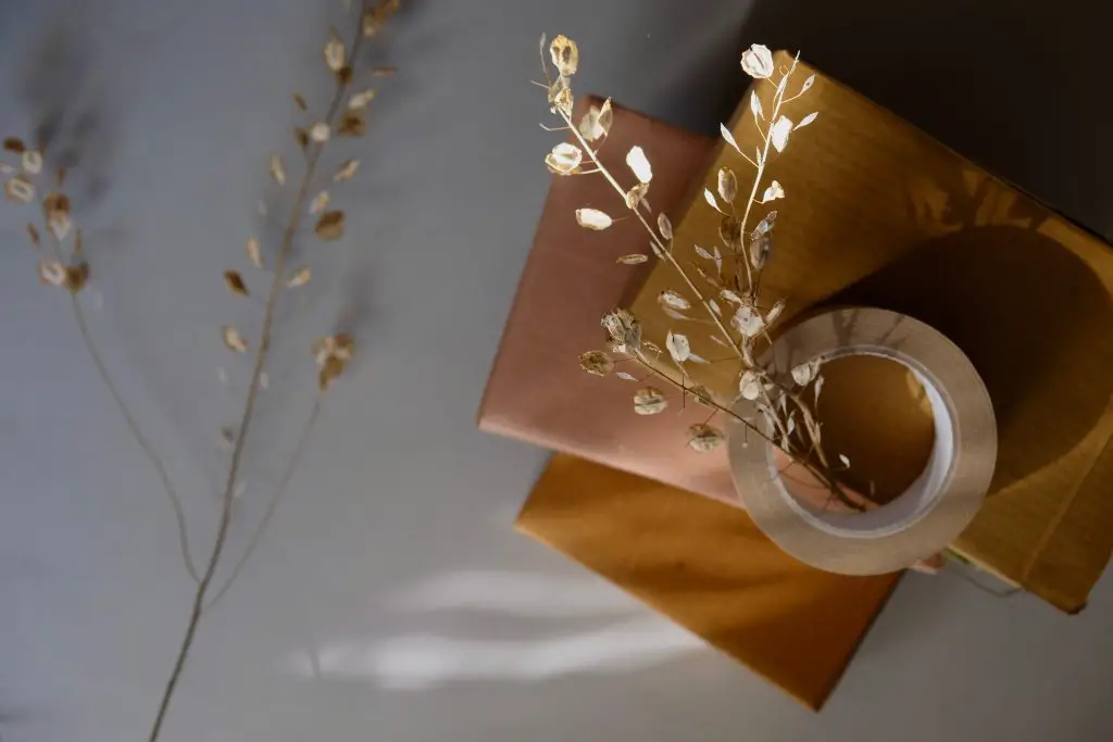 A pile of small packages wrapped in brown paper. On top of them is a roll of brown tape. There are sprigs of dried plants on top of the tape and next to the packages.