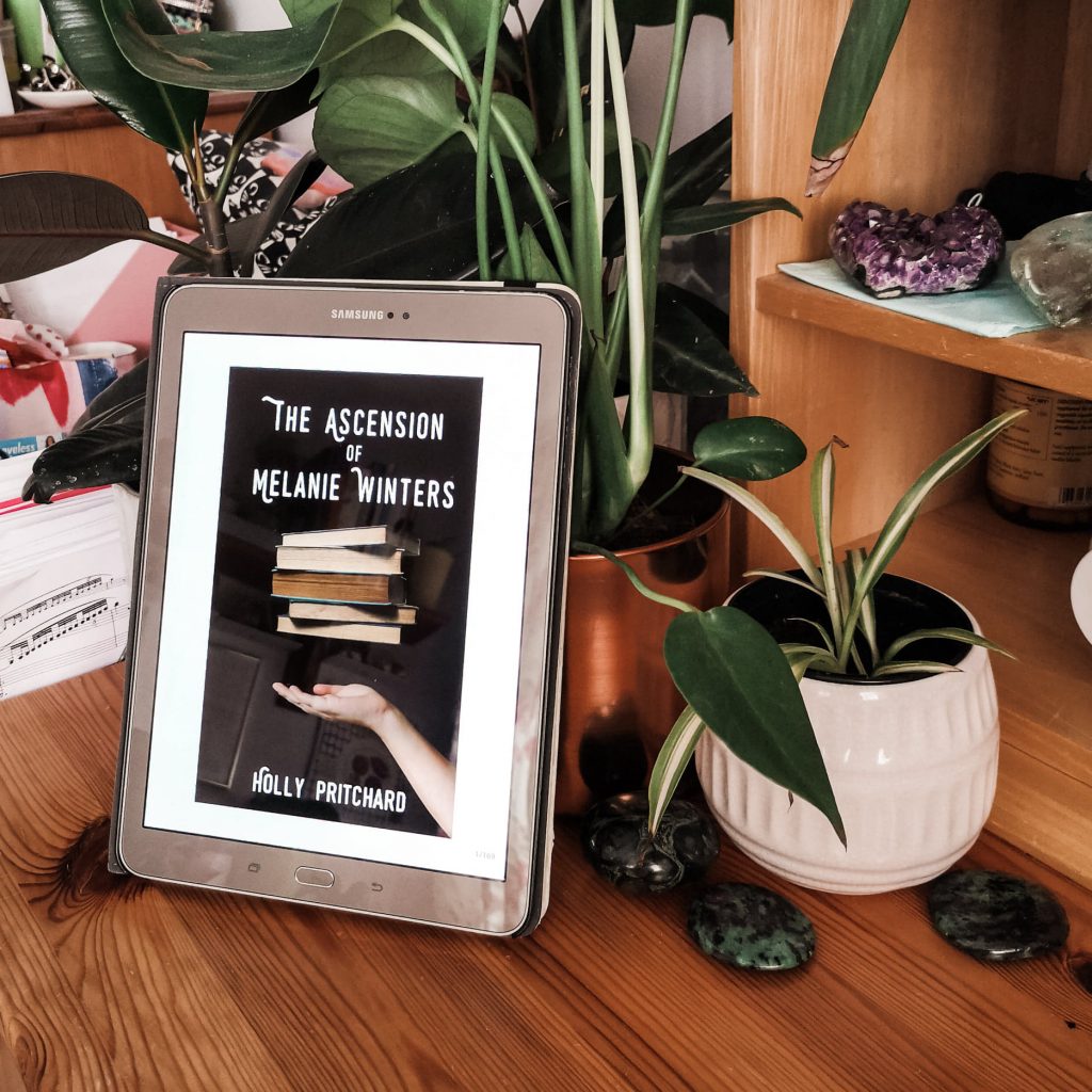The cover of 'The Ascension of Melanie Winters' shown on a tablet, which is propped up against some plant pots.