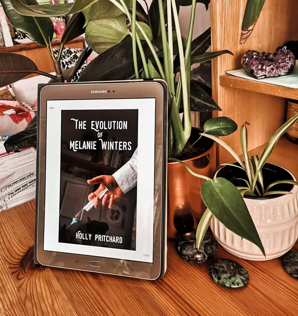 The cover of 'The Evolution of Melanie Winters' shown on a tablet, which is propped up against some plant pots.