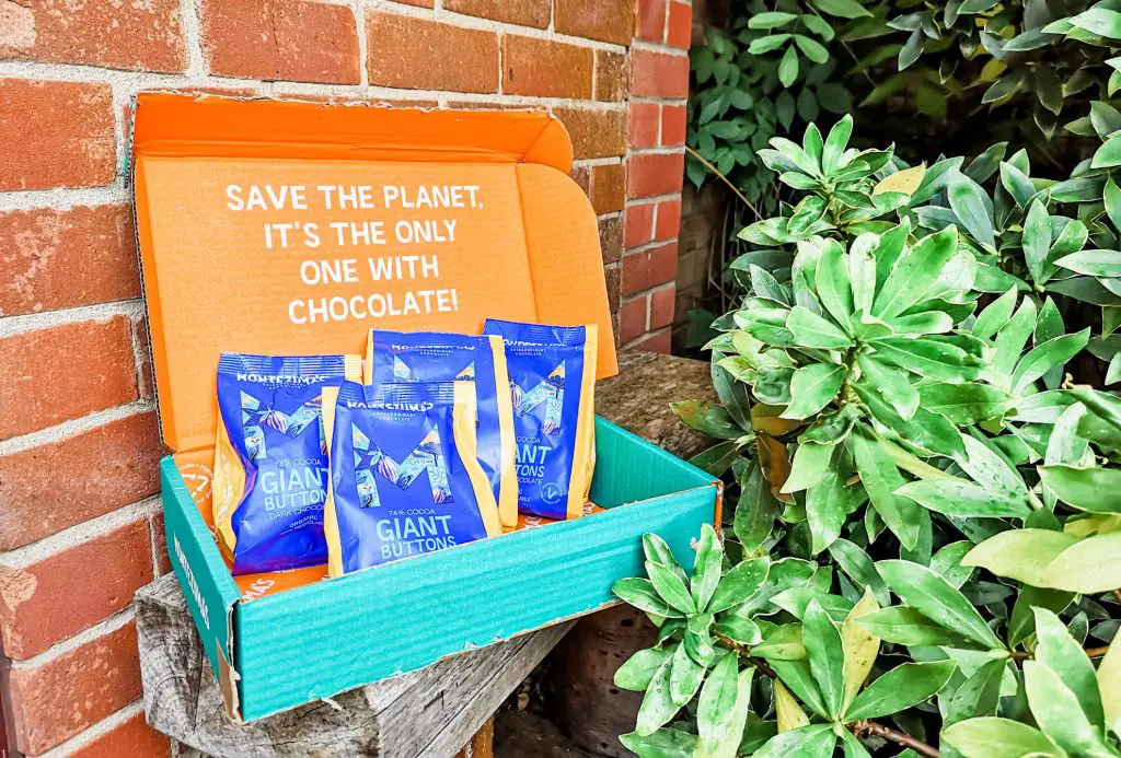 A colourful cardboard box sporting the phrase 'Save the planet, it's the only one with chocolate!'. In the box are 4 bags of Montezuma's 74% cocoa giant chocolate buttons.