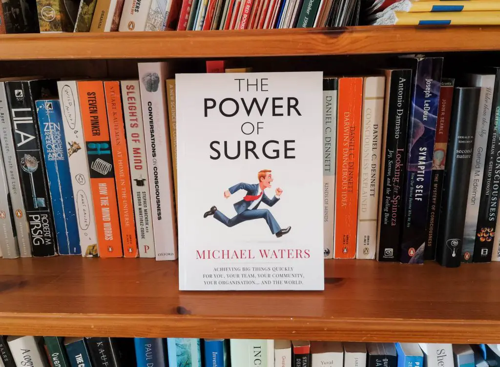 A paperback copy of The Power of Surge by Michael Waters, on a book shelf.