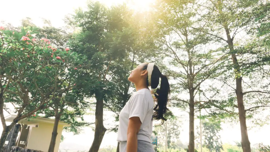 A woman standing outside, surrounded by trees and plants. The sun is shining, she is looking upwards and smiling.