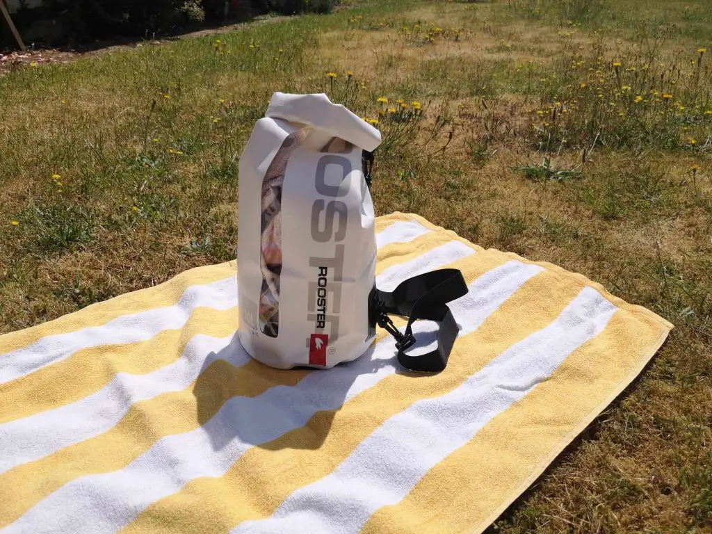 A white, Rooster brand dry bag on a stripy yellow and white towel which is laid out on a lawn.