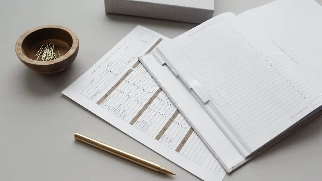 An open 'Finance Tracker' notebook, and a Monthly Budget sheet, along with a gold coloured pen and a wooden pot of paperclips.