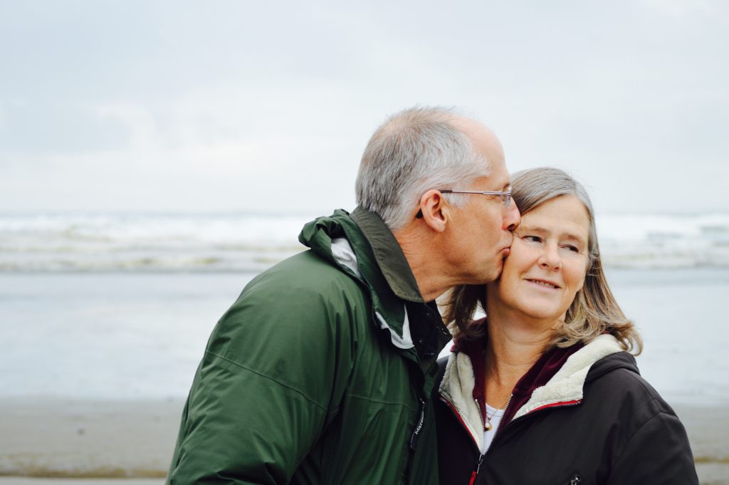 An older man kissing an older woman on the cheek. In the background is the sea.