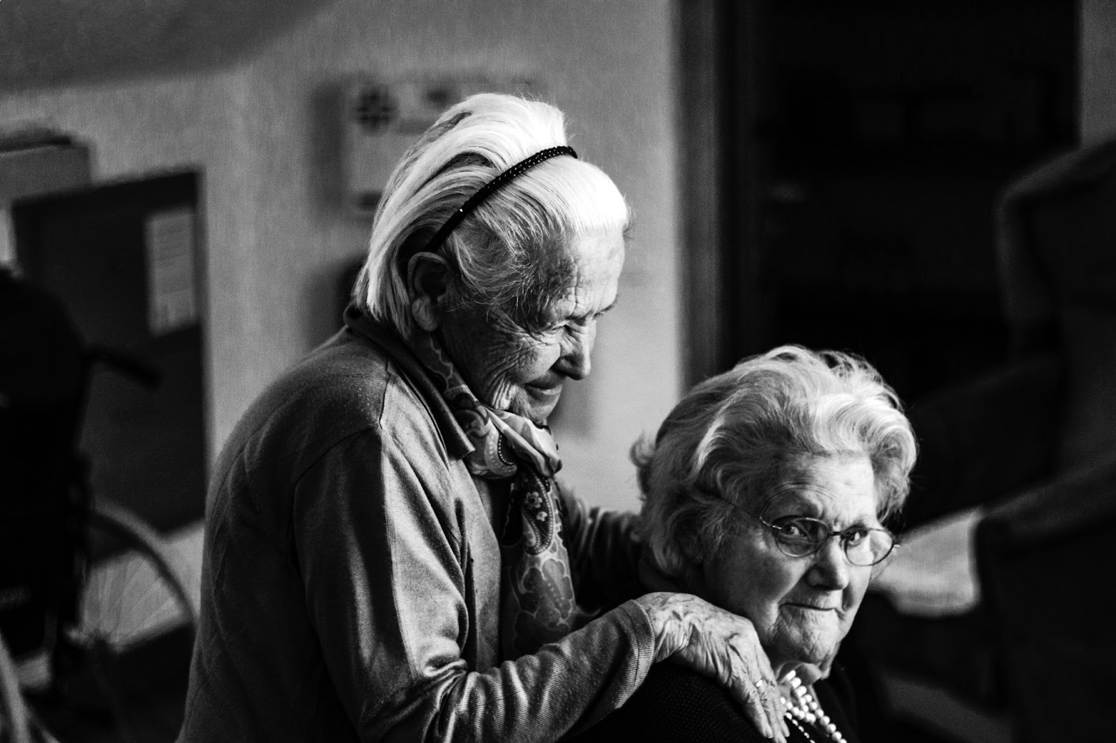 Two older women, one has her hands on the other's shoulders and is smiling.