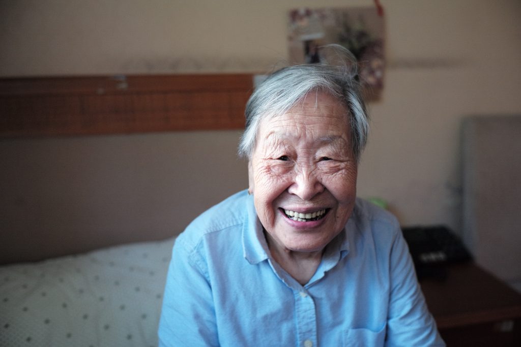 An older woman sitting on her bed and smiling.