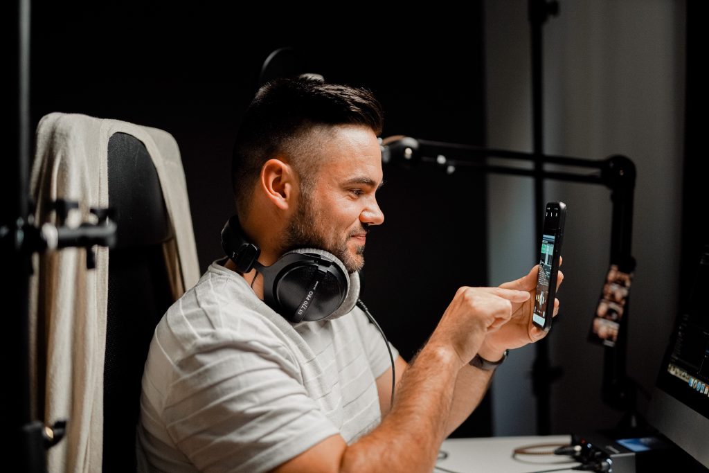 A man wearing headphones around his neck and surrounded by recording equipment. He is looking at his phone and smiling.