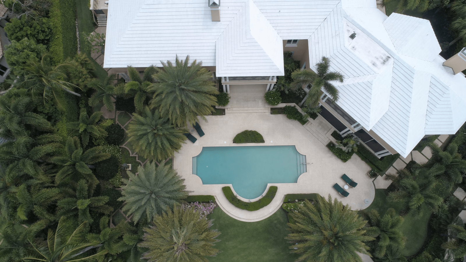 A birds-eye-view of a beautiful modern home with a white roof and a swimming pool, surrounded by tropical trees.