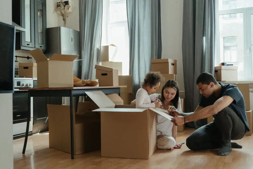 A family drawing on a cardboard box with marker pens. They are surrounded by other cardboard boxes.