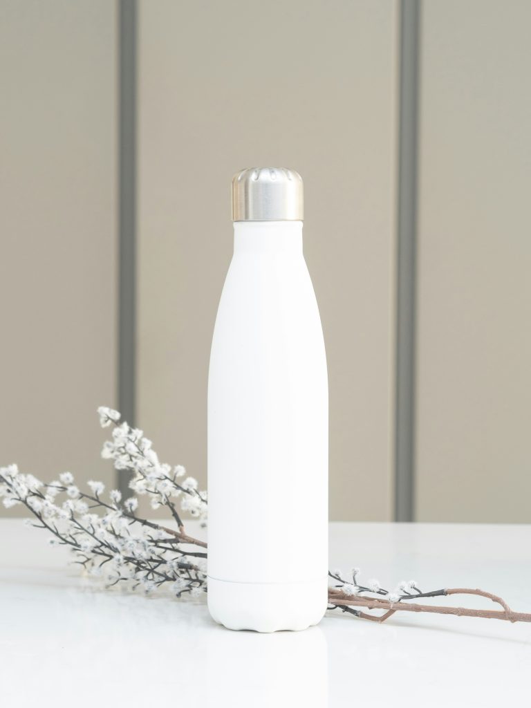 A white, metal water bottle on a table with a branch of blossom behind it.