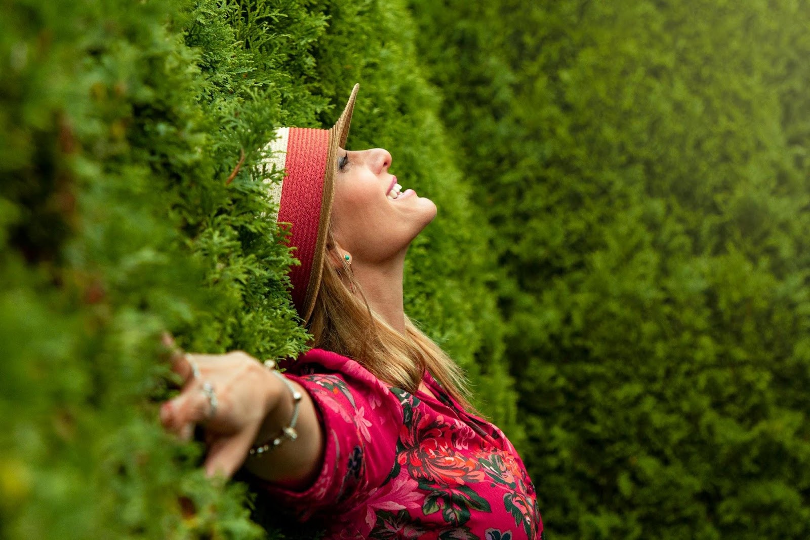 A women wearing a vibrant red top and a straw hat, leaning back against a hedge and smiling.