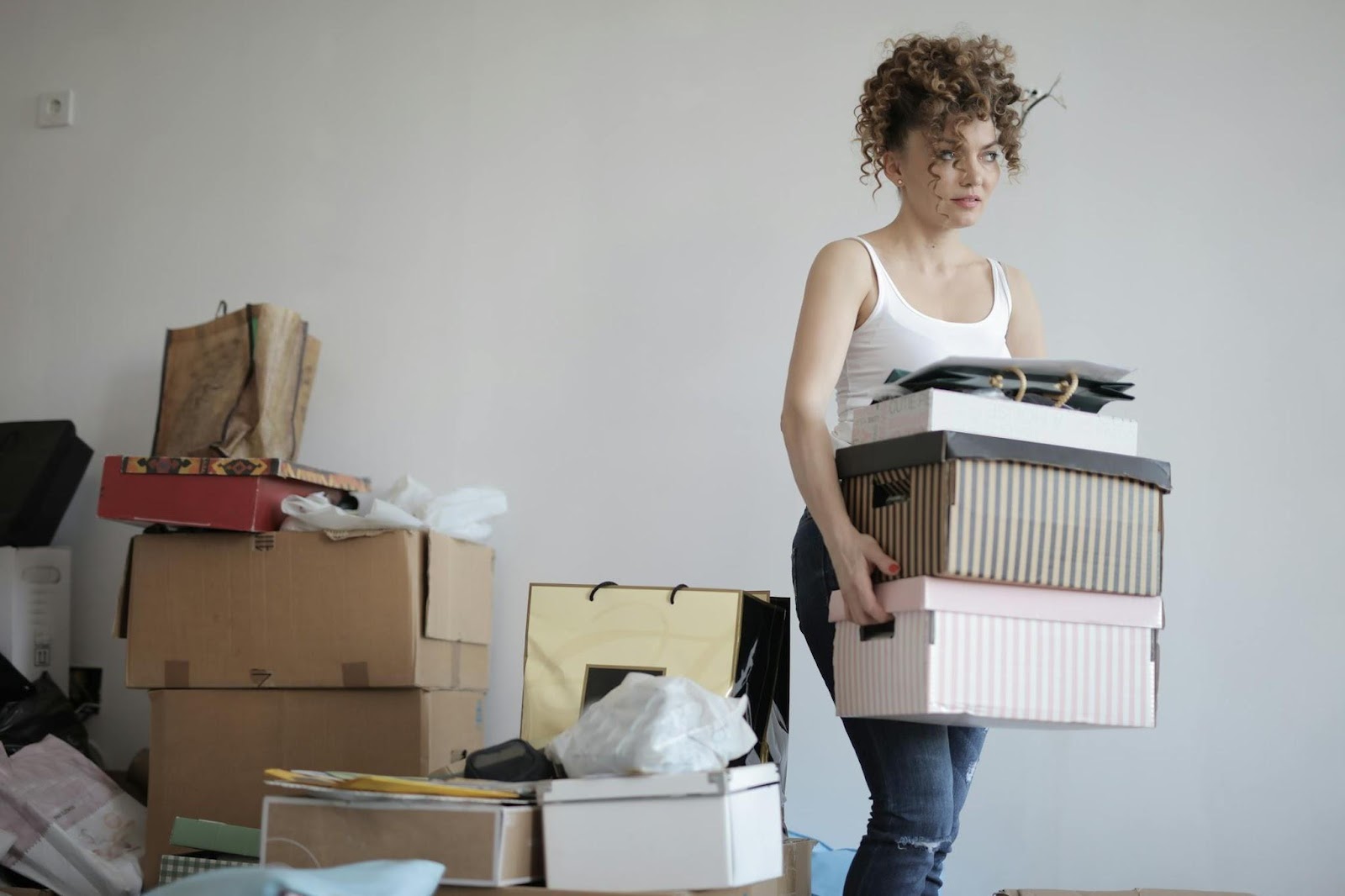 A woman carrying a stack of boxes out of a cluttered room.