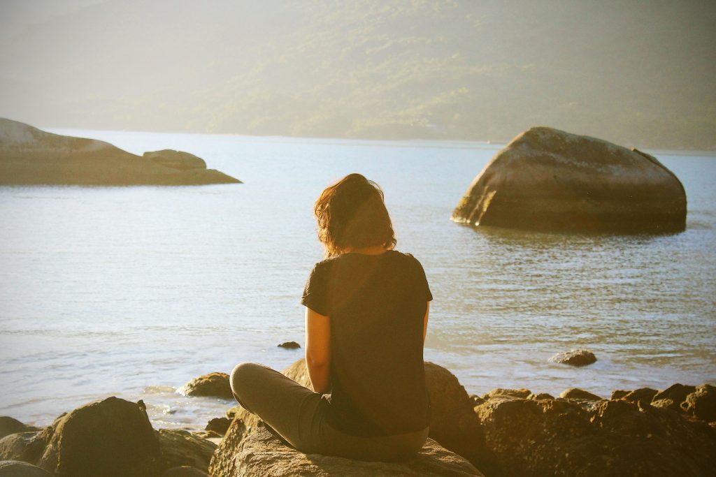 A woman sitting meditatively on a rock by a lake.