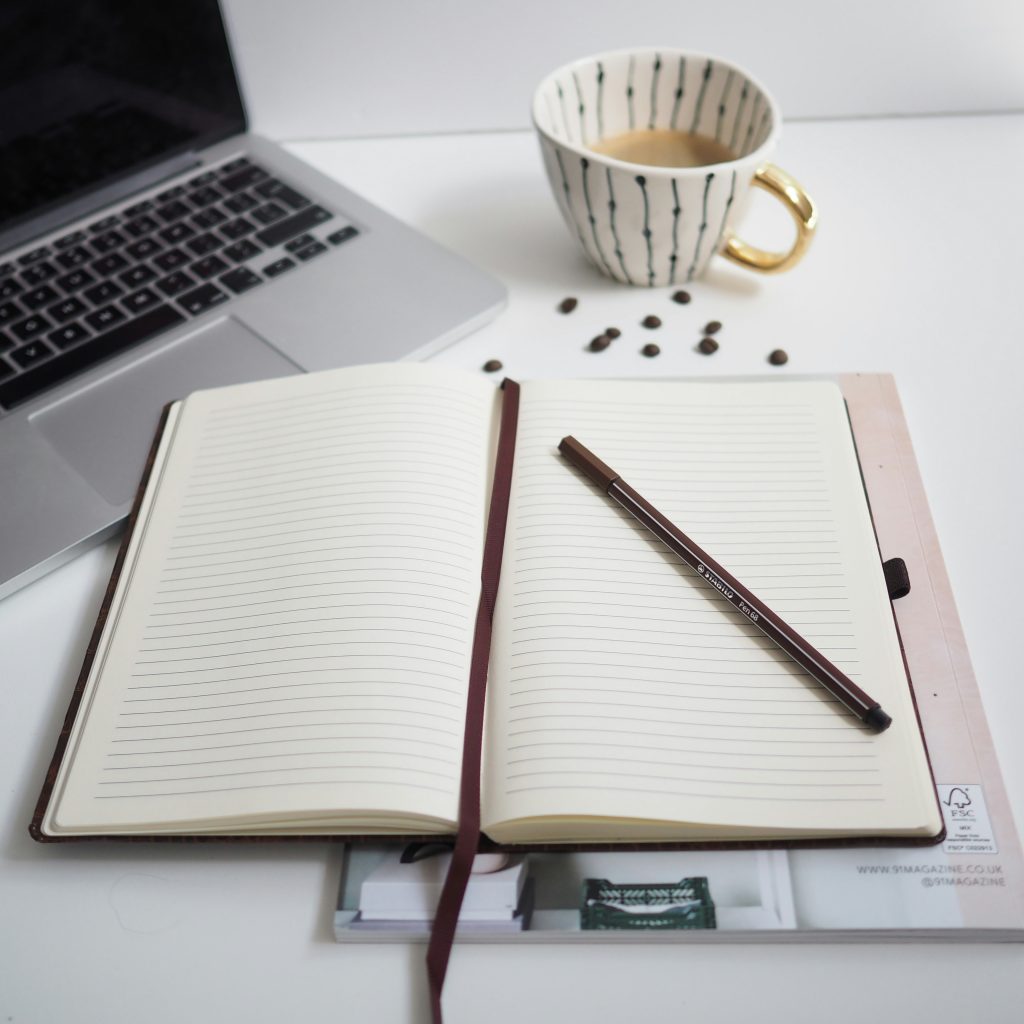An open notebook and pen, a cup of tea and a laptop on a desk.