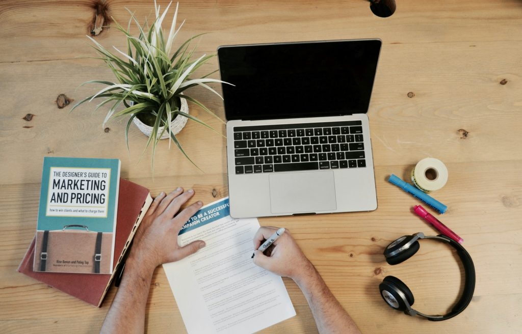 A birds-eye-view of a desk with an open laptop, several books, a potted plant, stationery and headphones.